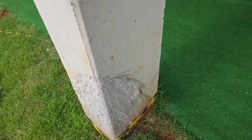 Base de pilar de concreto com trecho de textura e cor diferentes no canto inferior; piso gramado e fita amarela (quadra coberta). Origem do trecho: A CONFERIR.