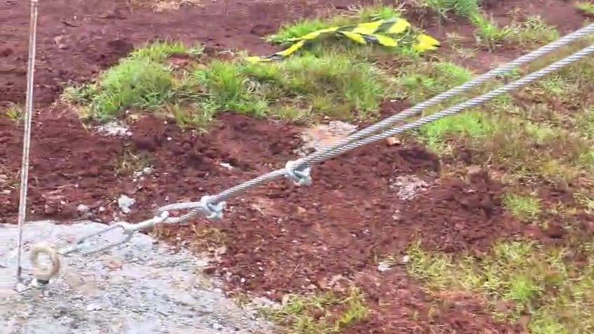 Par de cabos de aço com laços/grampos ligados a olhal em concreto no solo; fita de sinalização ao fundo (quadra coberta).