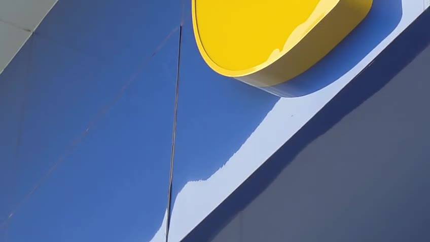 Painel ACM azul com junta/fresta vertical estreita junto a letreiro amarelo em relevo (Colégio).